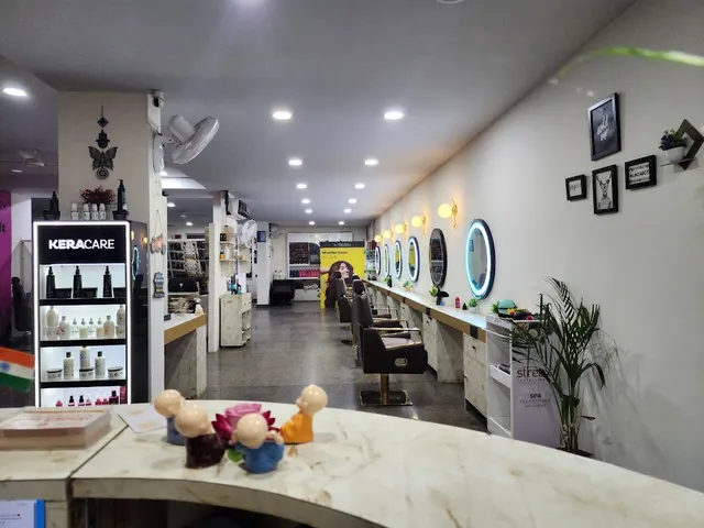 A row of styling stations along a mirrored wall, each with a bulb-lit round mirror, a leather chair and a marble-look counter. A lit display case of KeraCare hair care products stands in the foreground.